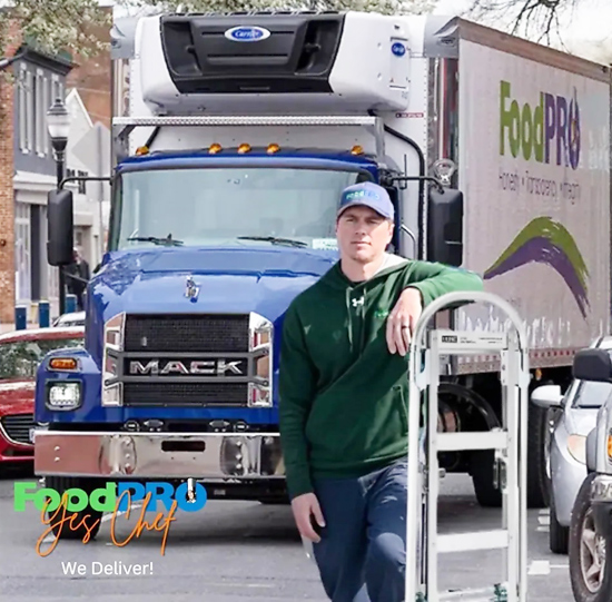 Blue Mack truck with FoodPRO trailer delivers food in a bustling urban neighborhood.