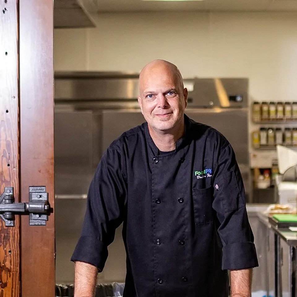 Chef in professional kitchen, smiling in stylish attire amidst stainless steel equipment and spices.